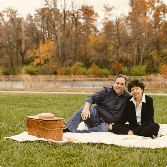 A joyful senior couple enjoying a picnic on a blanket in a park, surrounded by autumn foliage. They look happy and healthy, embracing a peaceful moment together.